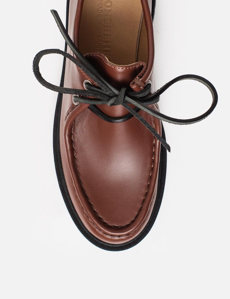 May include: A brown leather shoe with a moccasin-style toe, featuring black leather laces tied in a bow through silver-toned metal eyelets. The shoe has a thick black sole and a tan interior lining with the text ONCHENI MADE IN UKRAINE visible.