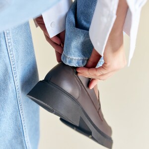 K&ouml;nnte beinhalten: Ein brauner Lederloafer mit einer dicken, schwarzen Sohle. Der Schuh wird angezogen, mit dem Aufschlag einer hellblauen Jeans und einem wei&szlig;en Hemds&auml;rmel sichtbar. Der Schuh scheint Gr&ouml;&szlig;e 42 zu haben.