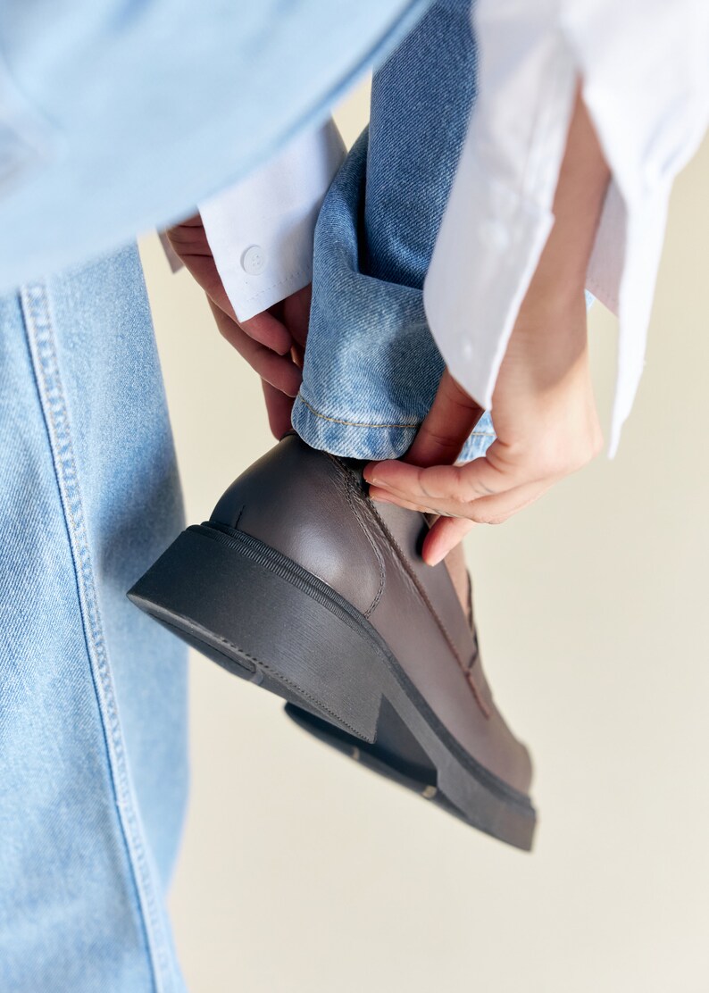 K&ouml;nnte beinhalten: Ein brauner Lederloafer mit einer dicken, schwarzen Sohle. Der Schuh wird angezogen, mit dem Aufschlag einer hellblauen Jeans und einem wei&szlig;en Hemds&auml;rmel sichtbar. Der Schuh scheint Gr&ouml;&szlig;e 42 zu haben.