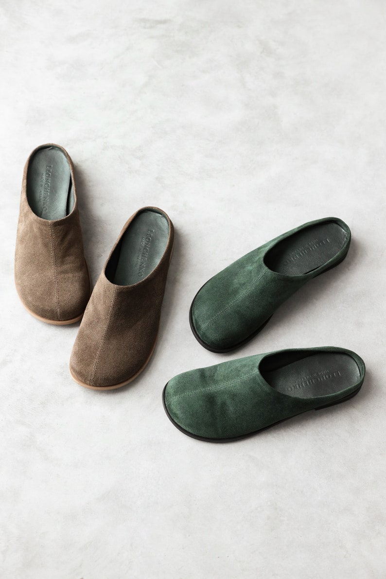 May include: Two pairs of slip-on mules in suede leather, one pair in brown and one pair in dark green. Each shoe features a centre seam, a rounded toe, and an open back. The interior footbed is stamped with the brand name LEONCHENKO.