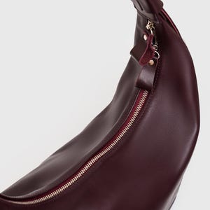 May include: A burgundy leather crossbody bag with a curved shape. The bag features a silver zipper and a matching burgundy interior. The strap is attached at the top and is also burgundy. The bag is made of a smooth, shiny leather.