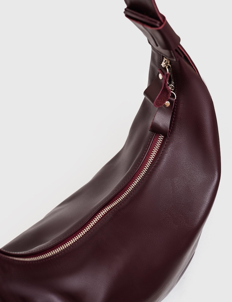 May include: A burgundy leather crossbody bag with a curved shape. The bag features a silver zipper and a matching burgundy interior. The strap is attached at the top and is also burgundy. The bag is made of a smooth, shiny leather.