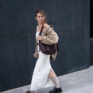 May include: A woman walking on a sidewalk, wearing a white dress, black shoes, and a tan jacket over a white tank top. She carries a large, dark burgundy shoulder bag. The background is a dark gray wall.