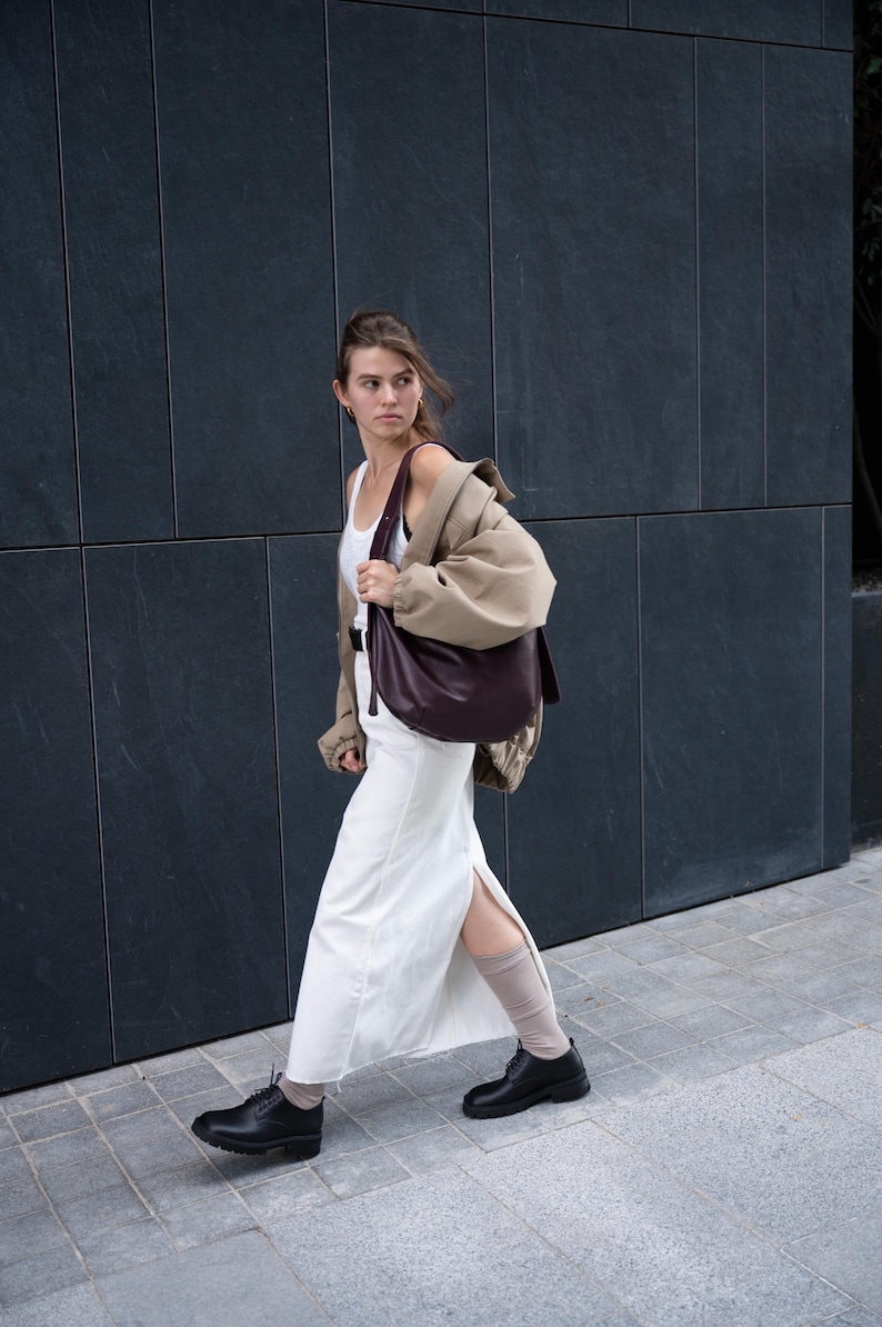 May include: A woman walking on a sidewalk, wearing a white dress, black shoes, and a tan jacket over a white tank top. She carries a large, dark burgundy shoulder bag. The background is a dark gray wall.
