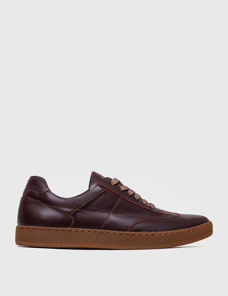 May include: A single brown leather sneaker with a stitched design and tan laces. The shoe has a brown sole and red stitching accents. The sneaker is a low-top style and is set against a white background.