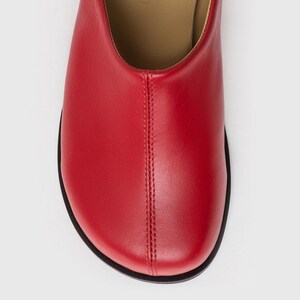 May include: Close-up of a red leather shoe with a rounded toe and a black sole. The shoe has a stitched seam down the center. The interior is a light tan color.