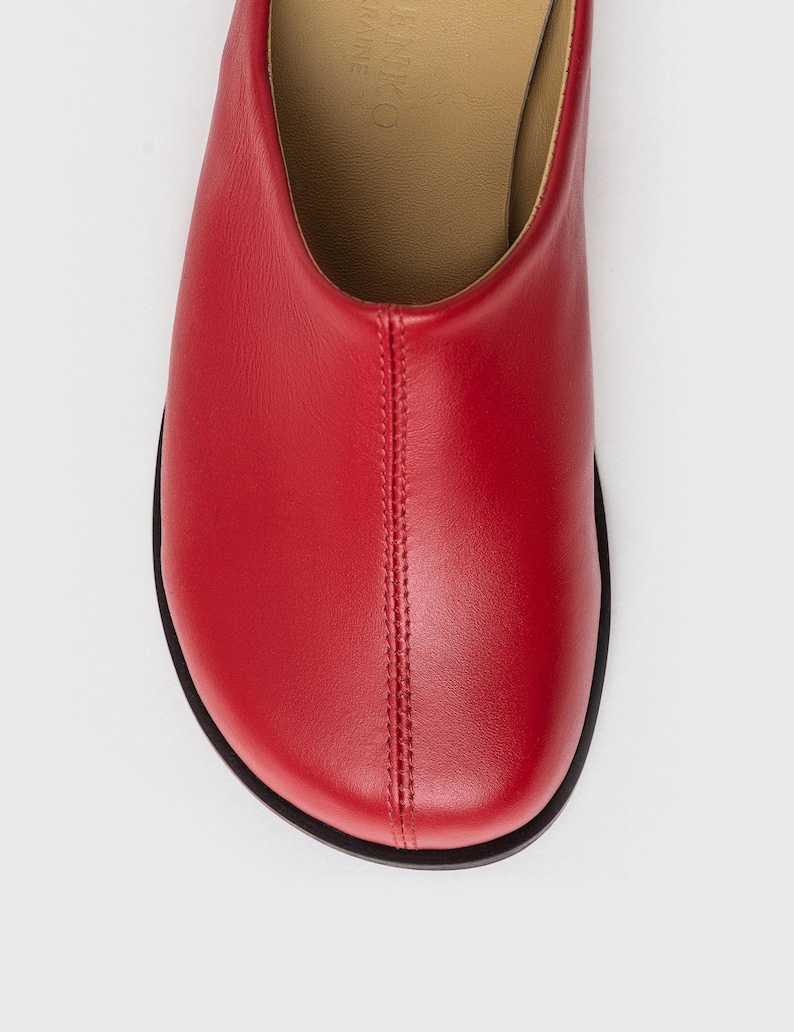 May include: Close-up of a red leather shoe with a rounded toe and a black sole. The shoe has a stitched seam down the center. The interior is a light tan color.