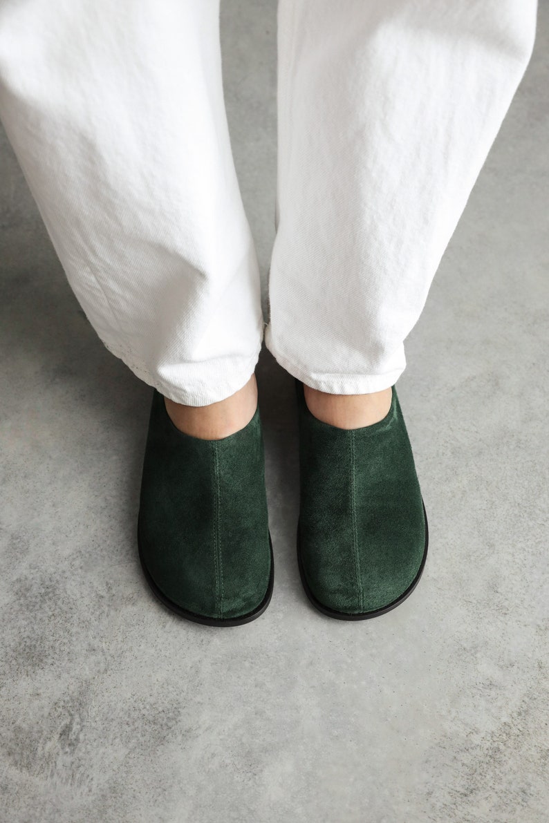 May include: A pair of dark forest green suede clogs featuring a rounded toe, a central vertical seam running down the front, and a black sole. The shoes are worn with white trousers, showcasing a simple slip-on design with a low-cut ankle opening.