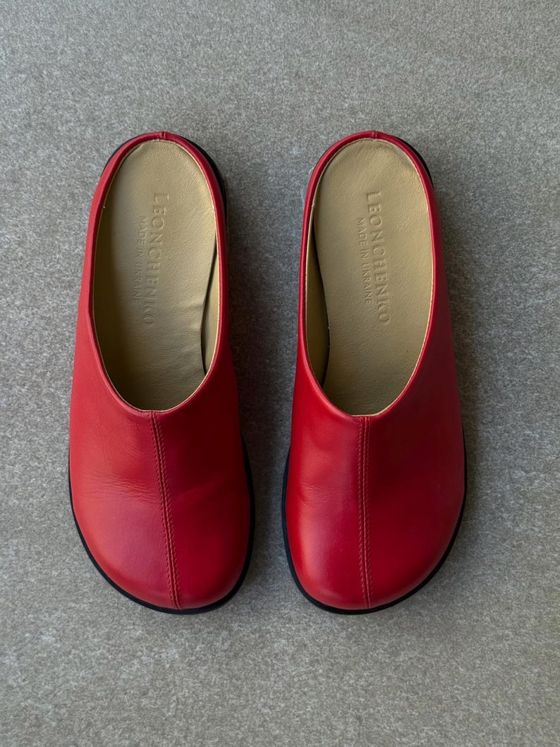 May include: A pair of red leather slip-on shoes with a rounded toe and a black sole. The interior is a light tan color, and the brand name "LEONCHENKO" is visible inside. The shoes are made in Ukraine.