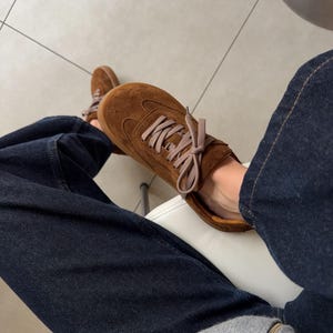 Puede incluir: Zapatillas de ante marr&oacute;n con cordones marr&oacute;n claro. Los zapatos se usan con vaqueros de mezclilla azul oscuro y una sudadera gris con cremallera. Las zapatillas tienen un dise&ntilde;o cl&aacute;sico y de perfil bajo.