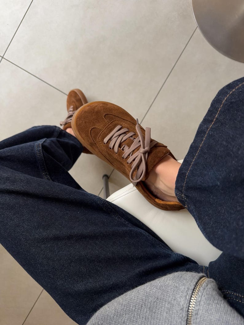 Puede incluir: Zapatillas de ante marr&oacute;n con cordones marr&oacute;n claro. Los zapatos se usan con vaqueros de mezclilla azul oscuro y una sudadera gris con cremallera. Las zapatillas tienen un dise&ntilde;o cl&aacute;sico y de perfil bajo.