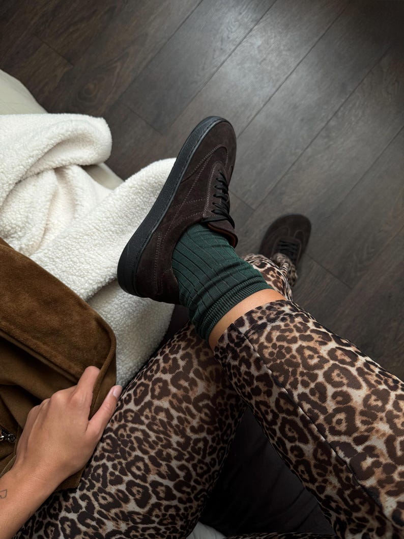 Puede incluir: Primer plano de una zapatilla de ante marr&oacute;n con suelas y cordones negros. El zapato se combina con calcetines verdes de canal&eacute; y leggings con estampado de leopardo. La imagen est&aacute; tomada desde arriba.