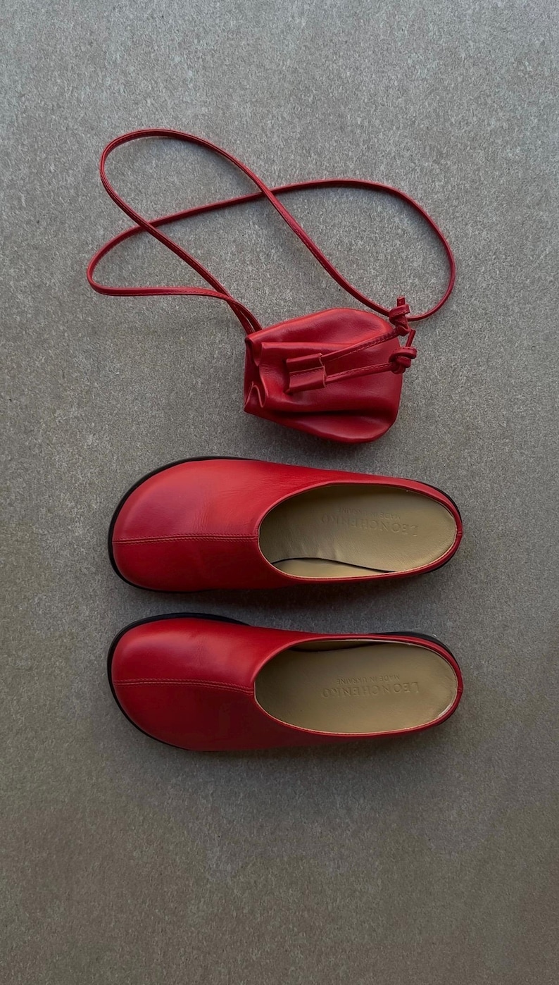 May include: A set of red leather footwear and a matching small purse. The footwear includes a pair of slip-on shoes with a rounded toe and a black sole. The purse is a small, drawstring style bag with a long strap.
