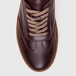 May include: Burgundy leather sneakers with tan laces and a brown sole. The shoes have a classic lace-up design with visible stitching details. The sole is approximately 1 inch thick.