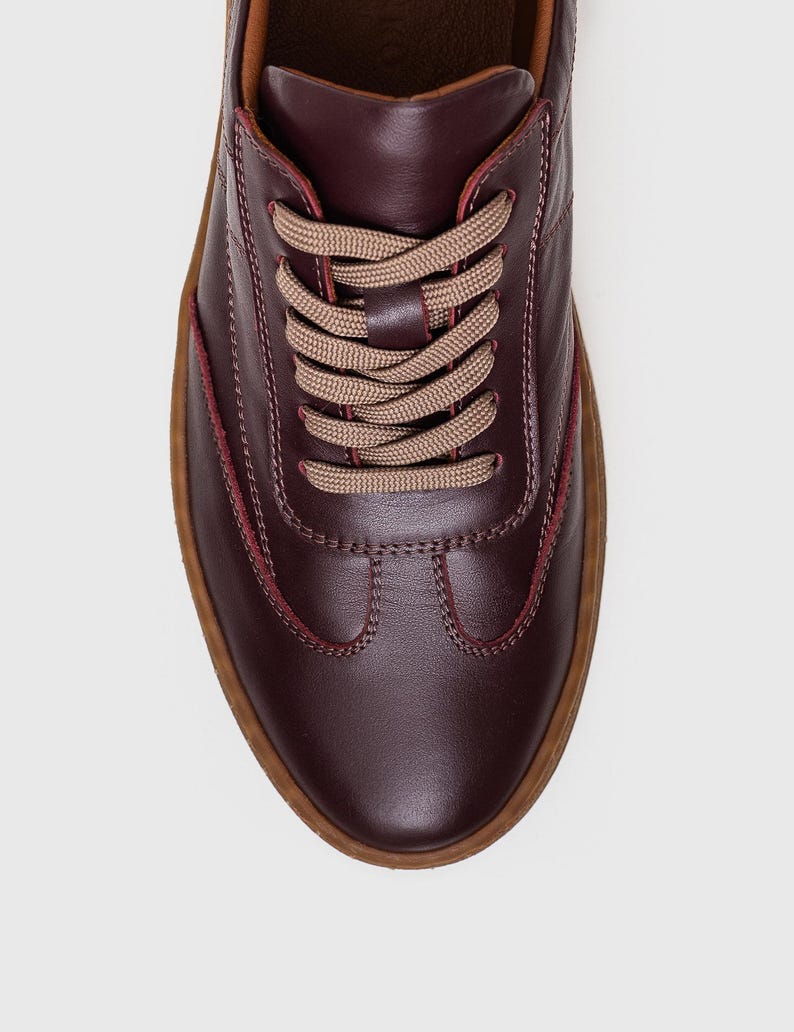 May include: Burgundy leather sneakers with tan laces and a brown sole. The shoes have a classic lace-up design with visible stitching details. The sole is approximately 1 inch thick.