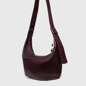 May include: A burgundy leather shoulder bag with a curved shape and a long, adjustable strap. The bag features a zipper closure and a minimalist design, suitable for everyday use.