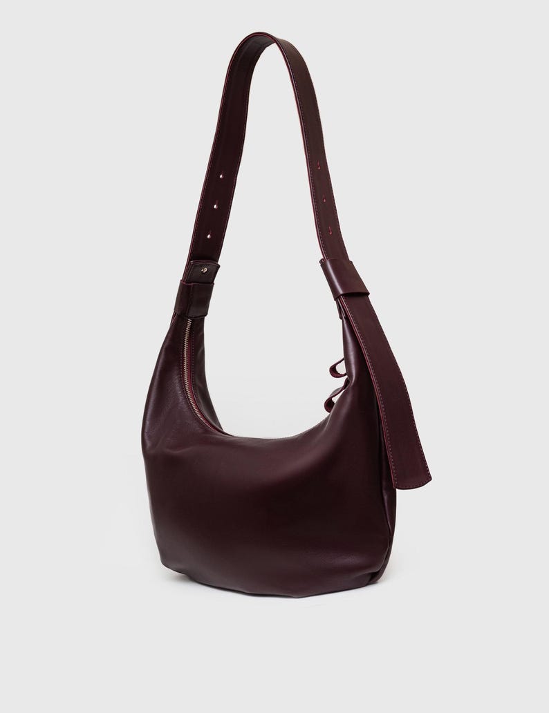 May include: A burgundy leather shoulder bag with a curved shape and a long, adjustable strap. The bag features a zipper closure and a minimalist design, suitable for everyday use.