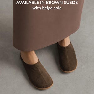 May include: Brown suede slip-on shoes with a rounded toe and a beige sole. The text on the image reads: AVAILABLE IN BROWN SUEDE with beige sole. These shoes feature a simple design with a centre seam running from the opening to the toe.