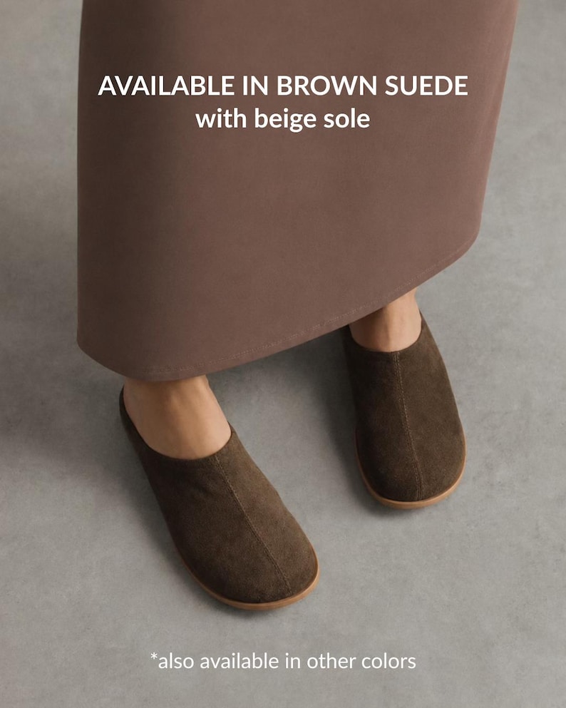 May include: Brown suede slip-on shoes with a rounded toe and a beige sole. The text on the image reads: AVAILABLE IN BROWN SUEDE with beige sole. These shoes feature a simple design with a centre seam running from the opening to the toe.