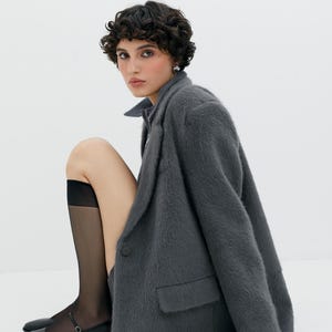 Puede incluir: Una mujer con un blazer gris peludo, medias negras y zapatos planos negros. Est&aacute; sentada con las piernas cruzadas y su mano derecha apoyada en el suelo.