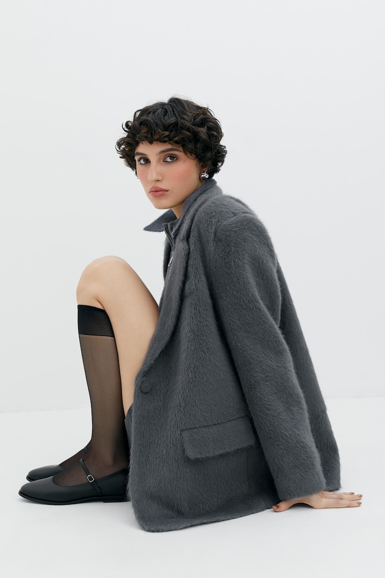 Puede incluir: Una mujer con un blazer gris peludo, medias negras y zapatos planos negros. Est&aacute; sentada con las piernas cruzadas y su mano derecha apoyada en el suelo.