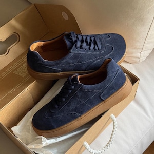 Puede incluir: Un par de zapatillas de ante azul marino con suelas y cordones marrones, presentadas en una caja de cart&oacute;n. Las zapatillas tienen un dise&ntilde;o cl&aacute;sico y bajo, adecuado para ropa informal. La caja est&aacute; abierta, revelando las zapatillas.