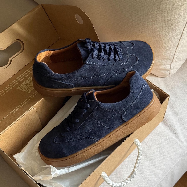 Puede incluir: Un par de zapatillas de ante azul marino con suelas y cordones marrones, presentadas en una caja de cart&oacute;n. Las zapatillas tienen un dise&ntilde;o cl&aacute;sico y bajo, adecuado para ropa informal. La caja est&aacute; abierta, revelando las zapatillas.
