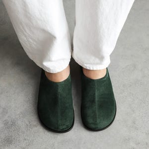 May include: A pair of dark forest green suede slip-on shoes with a rounded toe, a central vertical seam running down the front, and a thin black sole. The shoes are worn with white trousers, showcasing a simple, low-profile design.