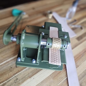 Puede incluir: Una máquina de trabajo de cuero de metal verde con un rodillo dorado y una tira de cuero que se alimenta a través de ella.