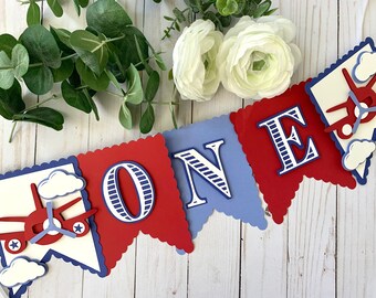 May include: A red, white, and blue paper banner with the word "ONE" in navy blue letters. The banner features airplanes and clouds, making it perfect for a first birthday party.