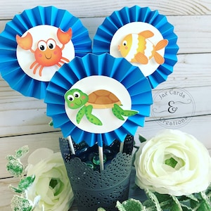 May include: Three blue paper fan decorations with white circles in the center. Each circle features a different sea creature: a crab, a fish, and a turtle. The decorations are arranged in a vase with green and blue glass beads. The text "Jax Cards & Creations" is visible in the background.