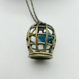 May include: A bronze-coloured birdcage pendant necklace with a blue bird inside. The cage has a detailed, openwork design and hangs from a delicate chain. The bird is a vibrant blue colour, contrasting with the bronze.