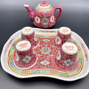 May include: A vintage Chinese tea set with a red teapot and four cups on a patterned tray. The set features a floral and geometric design in red, white, and gold. The cups and teapot have Chinese characters.