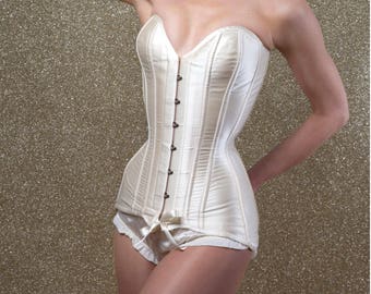 May include: A cream-colored corset with a sweetheart neckline and vertical boning. The corset features a series of small clasps down the front and is paired with matching high-waisted briefs. The background is a glittery gold.