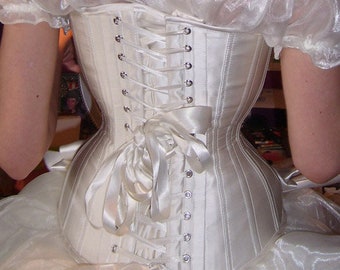 May include: A white satin corset with a lace-up back and a white ribbon bow. The corset has a series of metal eyelets and is worn over a white tulle skirt.