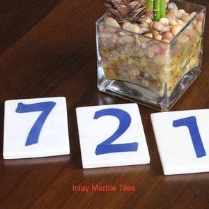 May include: Three white ceramic tiles with blue numbers 7, 2, and 1. The tiles are on a dark brown wooden surface. The text "Inlay Marble Tiles" is printed below the tiles.
