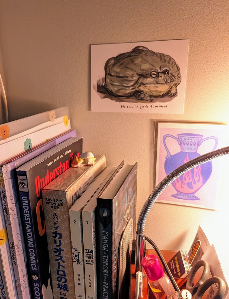 Pu&ograve; includere: Stampa a muro di una rana verde con la scritta "He too, is quite famished." Sotto, una stampa rosa di un vaso con fiamme blu e viola. Libri e una lampada da scrivania in primo piano.