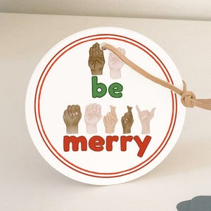 May include: White round ornament with red and green text that says "be merry" and features a row of hands signing the phrase in American Sign Language. The ornament has a brown string attached to it. A business card with the text "Sunday Table Simple + Playful Paper Goods" is in the background.