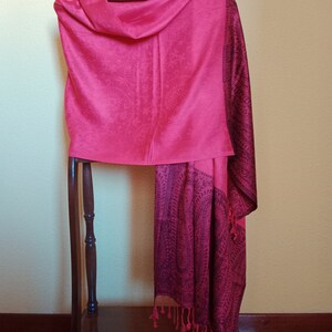 May include: A pink and black paisley patterned shawl with fringe hanging from the bottom edge. The shawl is draped over a wooden stand.