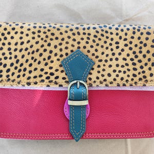 Puede incluir: Un bolso rectangular con solapa con estampado de guepardo y cuerpo rosa brillante. El bolso presenta un cierre de hebilla turquesa con una hebilla plateada. El bolso es de cuero.