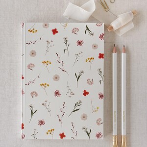 Könnte beinhalten: Ein weißes Notizbuch mit einem floralen Muster aus roten, gelben und rosa Blumen. Zwei weiße Bleistifte mit goldenen Spitzen und eine Rolle weißes Band liegen neben dem Notizbuch. Zwei goldene Büroklammern sind ebenfalls zu sehen.