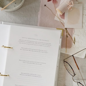 May include: A wedding planning binder with gold accents, open to a page with questions. The scene includes a lit candle, dried flowers, stationery, and a pair of glasses. The binder's questions are about wedding details.