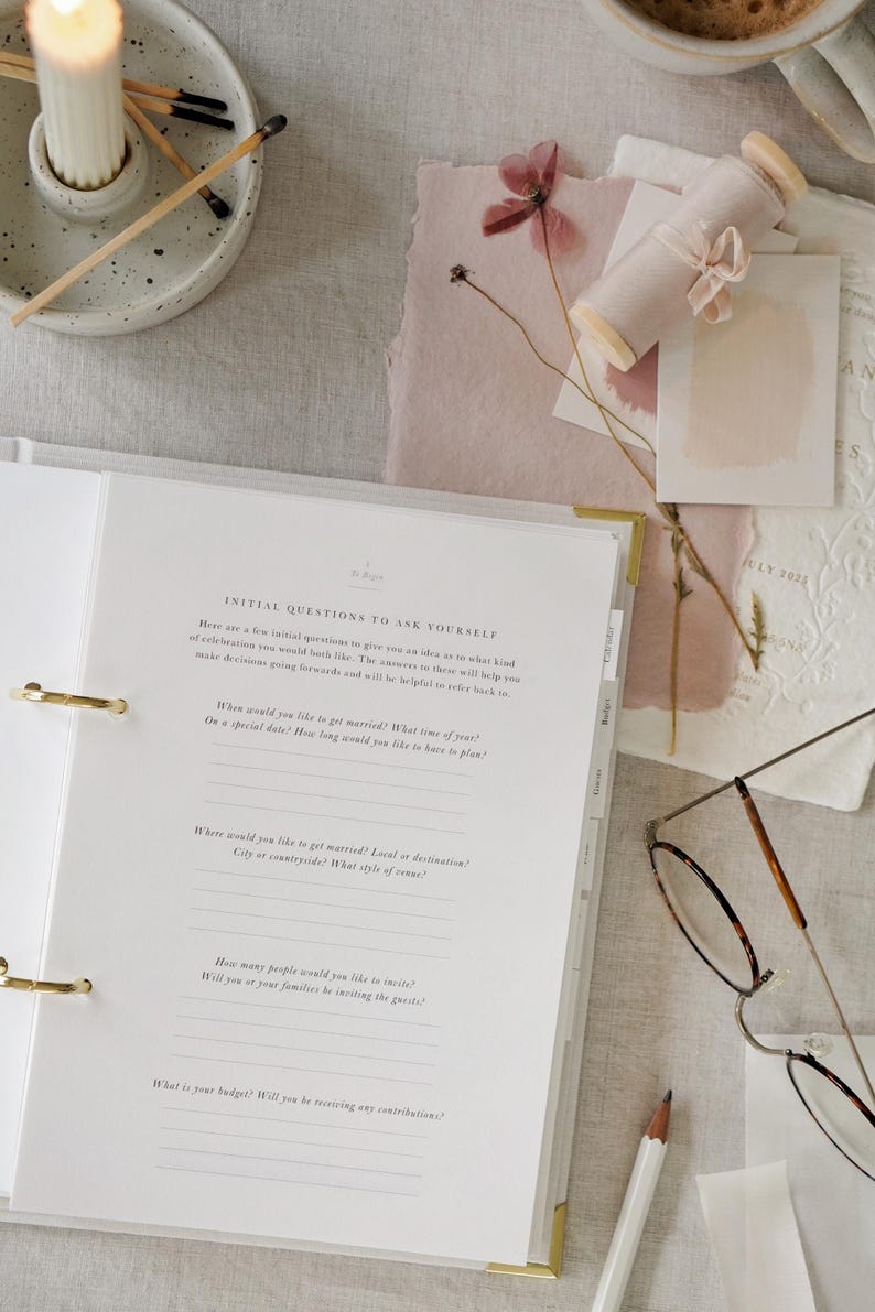 May include: A wedding planning binder with gold accents, open to a page with questions. The scene includes a lit candle, dried flowers, stationery, and a pair of glasses. The binder's questions are about wedding details.