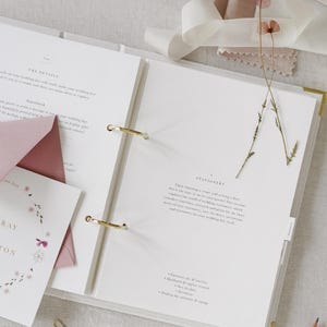 May include: An open white wedding planner with gold accents, featuring pages with text and gold ring binders. Accompanying stationery includes a pink envelope, invitation cards with floral designs, and a pencil. A cup of coffee and dried flowers are also present.