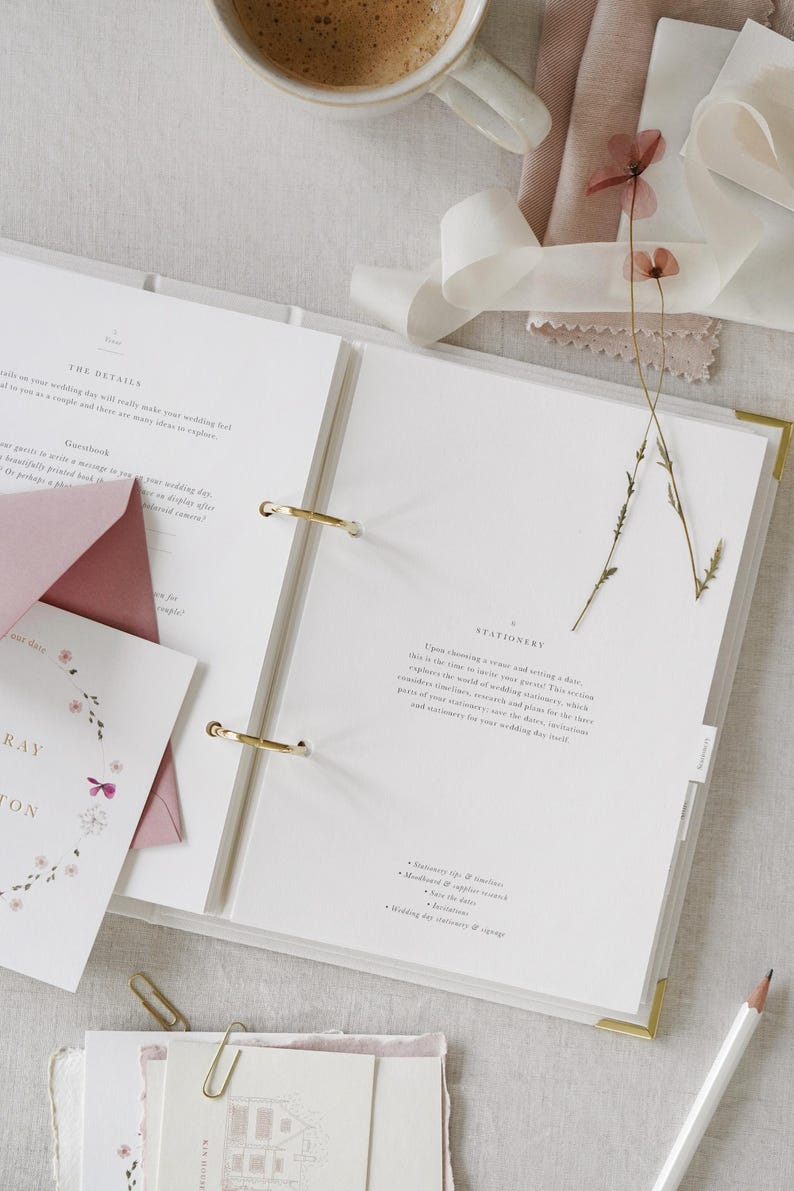 May include: An open white wedding planner with gold accents, featuring pages with text and gold ring binders. Accompanying stationery includes a pink envelope, invitation cards with floral designs, and a pencil. A cup of coffee and dried flowers are also present.