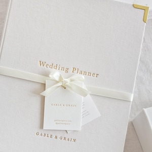 May include: A white linen-textured wedding planner with gold lettering and corner accents. The words "Wedding Planner" are printed on the cover. A white ribbon and tag with the brand name are tied around the planner.