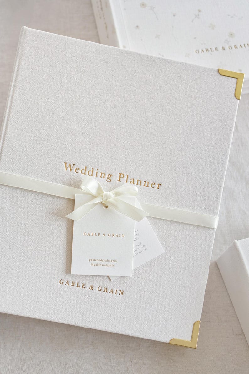 May include: A white linen-textured wedding planner with gold lettering and corner accents. The words "Wedding Planner" are printed on the cover. A white ribbon and tag with the brand name are tied around the planner.
