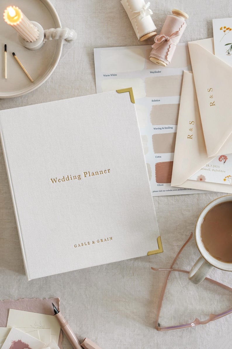 May include: A wedding planner book with gold lettering and corner protectors, alongside paint swatches, envelopes, and a cup of coffee. The book reads "Wedding Planner" and "GABLE & GRAIN". A lit candle and matches are also visible.