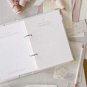 May include: An open white binder with gold accents, lined pages, and the text "INITIAL NOTES & IDEAS." The image includes spools of pink ribbon, stationery, a pencil, and a cup of coffee, suggesting a wedding planning theme.