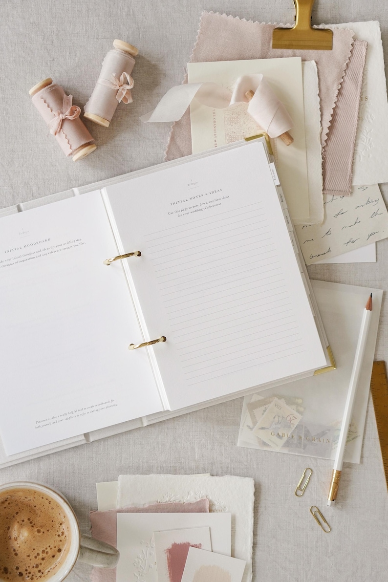 May include: An open white binder with gold accents, lined pages, and the text "INITIAL NOTES & IDEAS." The image includes spools of pink ribbon, stationery, a pencil, and a cup of coffee, suggesting a wedding planning theme.